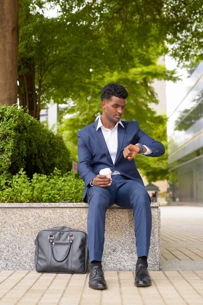 Retrato de um empresário africano verificando o tempo no relógio de pulso e segurando uma xícara de café para levar ao ar livre na cidade.