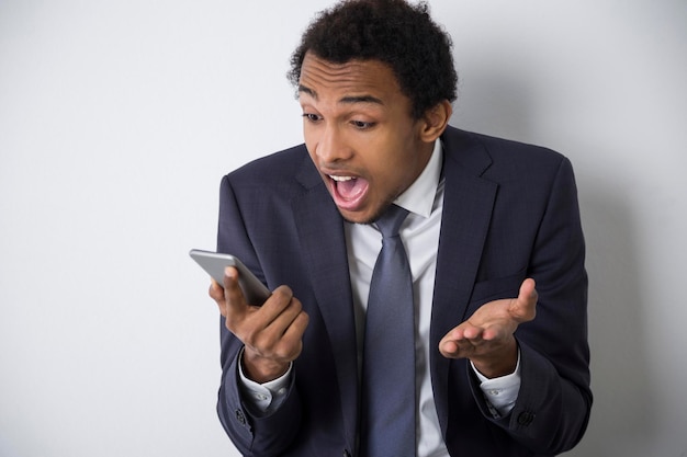 Retrato de um empresário afro-americano surpreso olhando para a tela do telefone inteligente em descrença. Conceito de emoções fortes