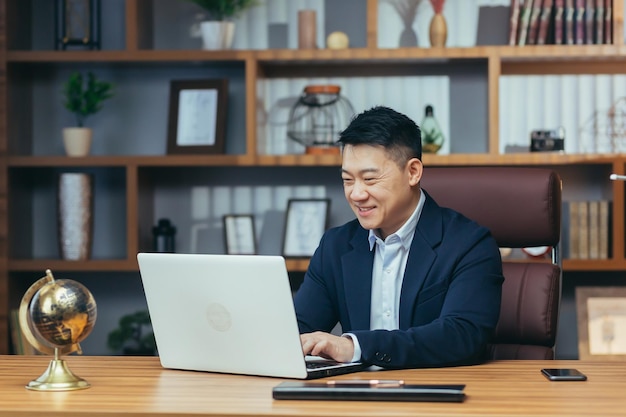 Retrato de um empresário asiático de sucesso em um escritório clássico trabalhando em um laptop sorrindo