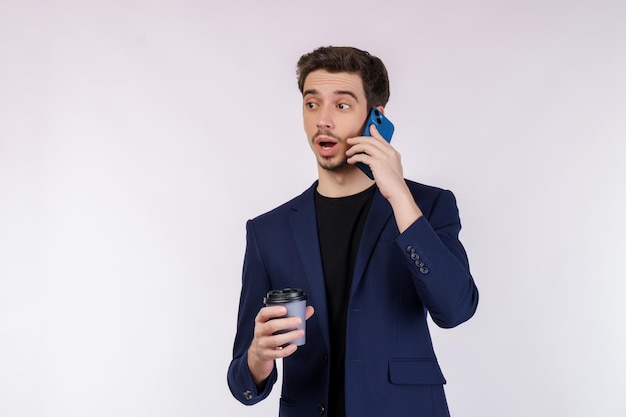 Retrato de um empresário bonitão chocado falando pelo celular e segurando café quente isolado sobre fundo branco