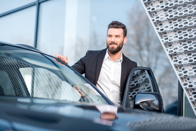 Retrato de um empresário bonitão em pé perto do carro ao ar livre, perto da fachada do edifício moderno