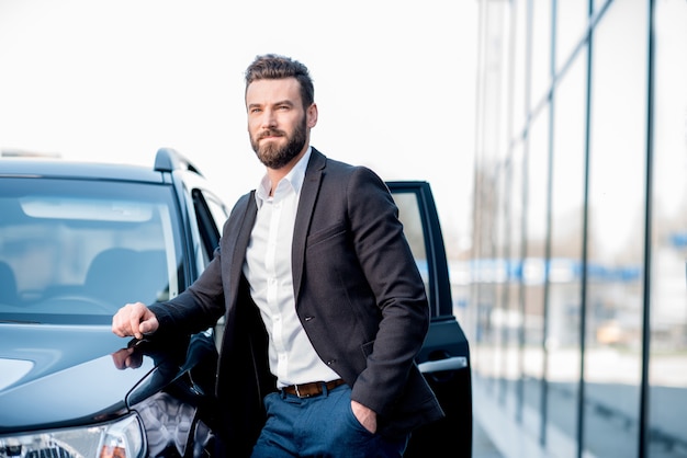 Retrato de um empresário bonitão em pé perto do carro ao ar livre, perto da fachada do edifício moderno