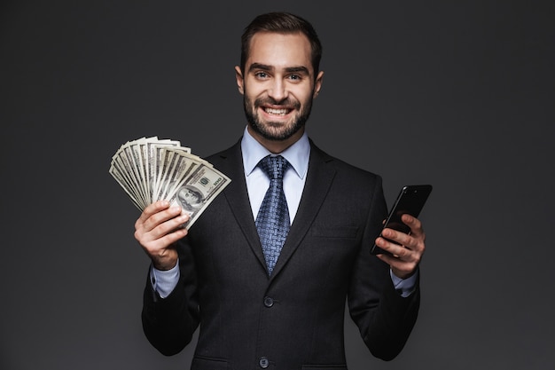 Retrato de um empresário bonito e confiante vestindo terno isolado, mostrando notas de dinheiro, usando telefone celular