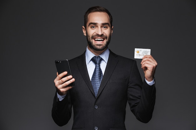 Retrato de um empresário bonito e confiante vestindo terno isolado, usando telefone celular, mostrando cartão de crédito de plástico