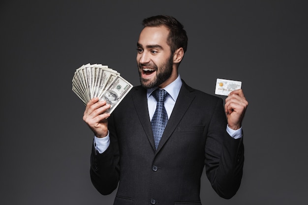 Retrato de um empresário bonito e confiante vestindo um terno isolado, mostrando notas de dinheiro, segurando um cartão de crédito de plástico