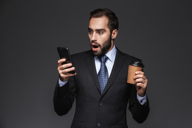 Retrato de um empresário chocado e confiante vestindo terno isolado, segurando uma xícara para viagem, usando telefone celular