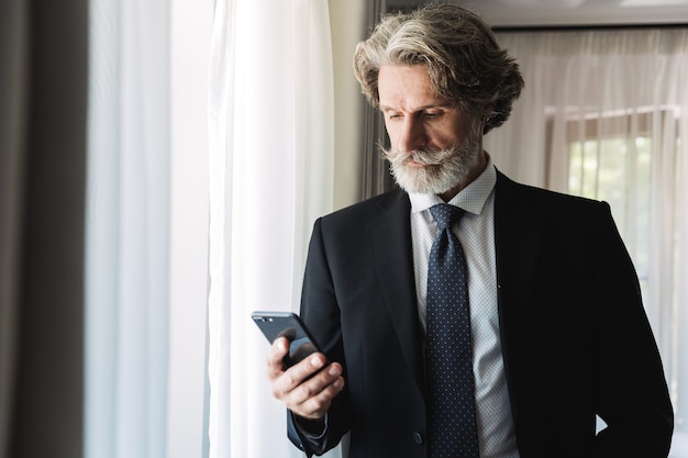 Retrato de um empresário de cabelos grisalhos barbudo maduro bonito posando dentro de casa em casa, perto da janela, vestido com roupas formais, usando telefone celular.