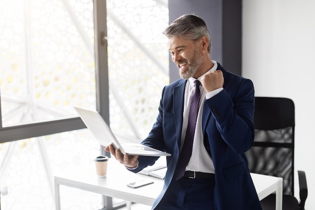 Retrato de um empresário de meia-idade feliz comemorando o sucesso com o laptop no escritório