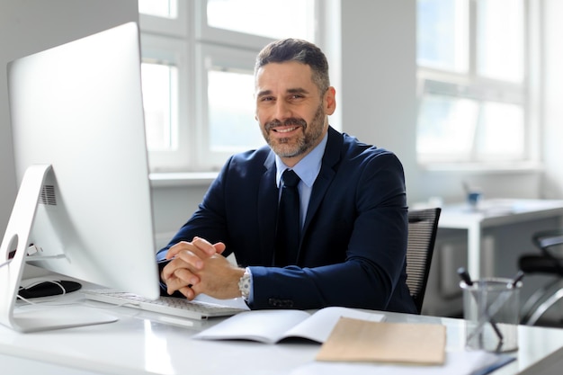 Retrato de um empresário de meia-idade feliz sentado em frente ao computador no local de trabalho no escritório