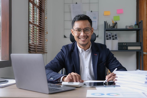 Retrato de um empresário do sexo masculino mostrando um rosto sorridente feliz como ele investiu com sucesso seu negócio usando computadores e documentos de orçamento financeiro no trabalho