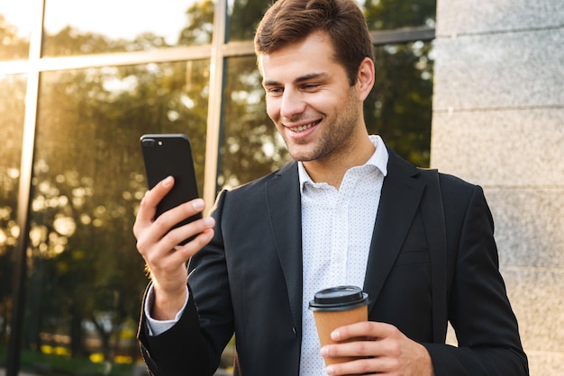 Retrato de um empresário executivo de terno segurando um telefone celular, ao ar livre perto do prédio com café para viagem