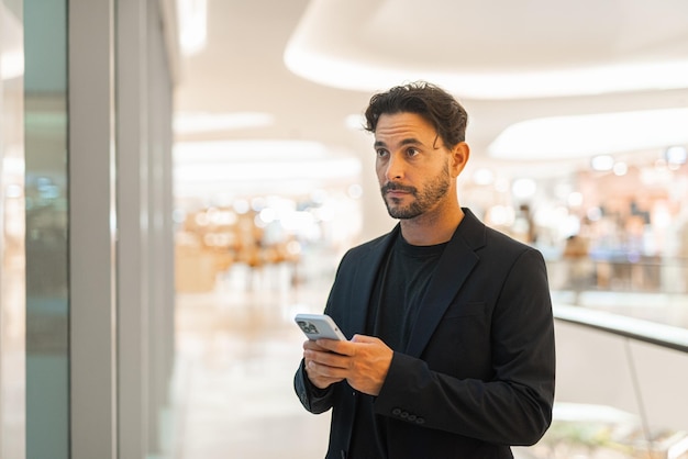 Retrato de um empresário hispânico bonito e feliz usando o telefone ao lado da janela no shopping