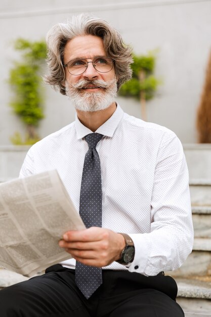 Retrato de um empresário idoso feliz em óculos segurando um jornal enquanto está sentado ao ar livre