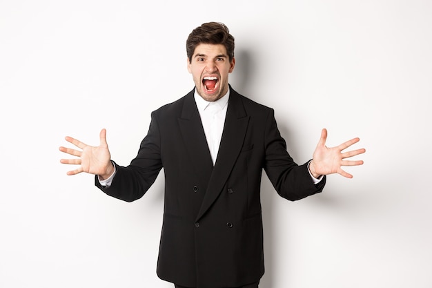 Retrato de um empresário irritado e frustrado de terno, gritando com raiva e apertando as mãos, em pé angustiado contra um fundo branco