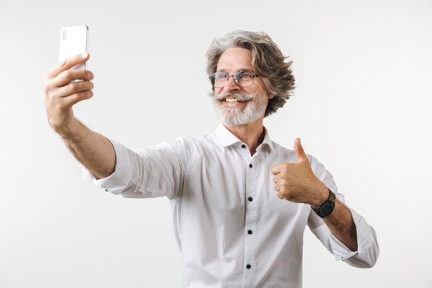 Retrato de um empresário maduro e bonito, vestido com roupa formal, em pé, isolado sobre uma parede branca, tirando uma selfie e dando os polegares para cima