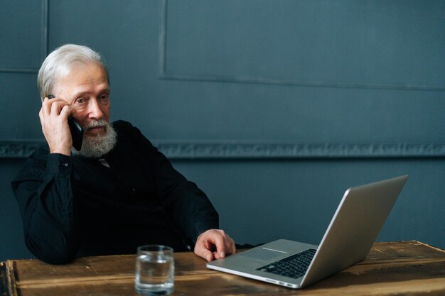 Retrato de um empresário mais velho maduro de cabelos grisalhos falando no celular sentado à mesa com o computador portátil