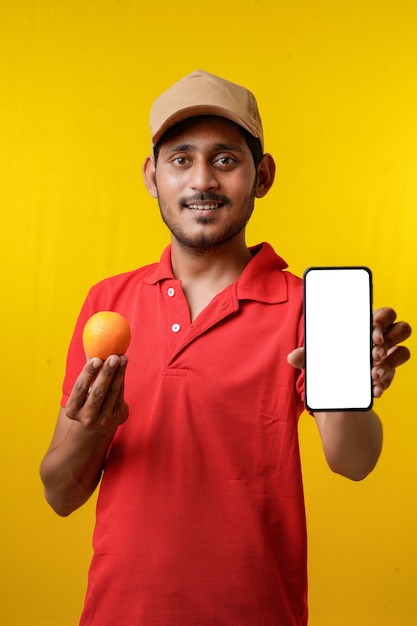 Retrato de um entregador de jovem feliz animado em camiseta vermelha e apresentando smartphone sobre fundo amarelo.