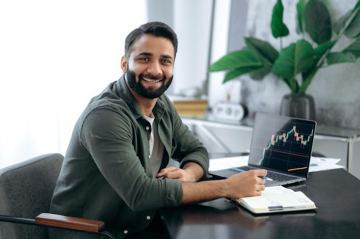 Foto retrato de um especialista em criptomoedas, corretor de bolsa de valores indiano ou árabe, bem-sucedido, atraente e positivo, sentado em uma mesa no escritório moderno, olhando para a câmera sorrindo amigável