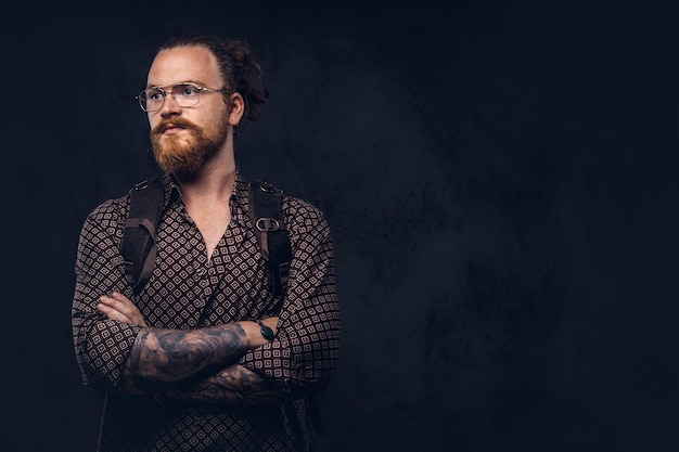 Retrato de um estudante hipster ruiva de óculos vestido com uma camisa marrom, segura uma mochila, posando em um estúdio. Isolado em um fundo escuro.