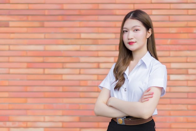 Retrato de um estudante tailandês adulto com uniforme de estudante universitário. Menina asiática bonita em pé com os braços cruzados sobre um fundo de tijolo.