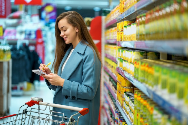 Retrato de um feliz sorridente atraente jovem comprador feminino com carrinho no corredor da loja com a lista de compras durante a compra de alimentos