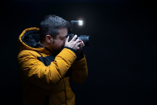 Retrato de um fotógrafo em um casaco de inverno amarelo tirando uma foto com flash com fundo preto