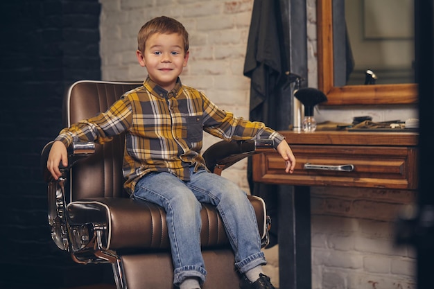 Retrato de um garotinho estiloso, vestido com camiseta e calça jeans, sentado em uma cadeira contra o local de trabalho do barbeiro, ele sorri e posa para a câmera