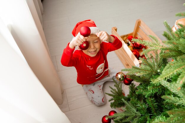 retrato de um garotinho fofo em um pijama vermelho decorando uma árvore de Natal conceito de ano novo de inverno