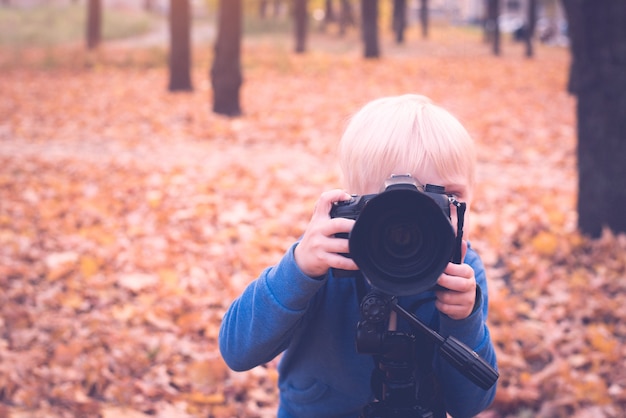 Retrato de um garotinho loiro com uma grande câmera dslr. Sessão de fotos no parque de outono