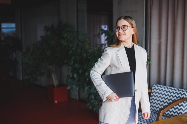 Retrato de um gerente de sucesso no escritório sorrindo durante o dia de trabalho mulher feliz com laptop aproveitando o tempo no escritório moderno Mulher loira em uma jaqueta bege e óculos