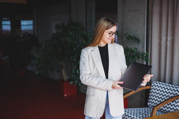 Retrato de um gerente de sucesso no escritório sorrindo durante o dia de trabalho mulher feliz com laptop aproveitando o tempo no escritório moderno mulher loira em uma jaqueta bege e óculos