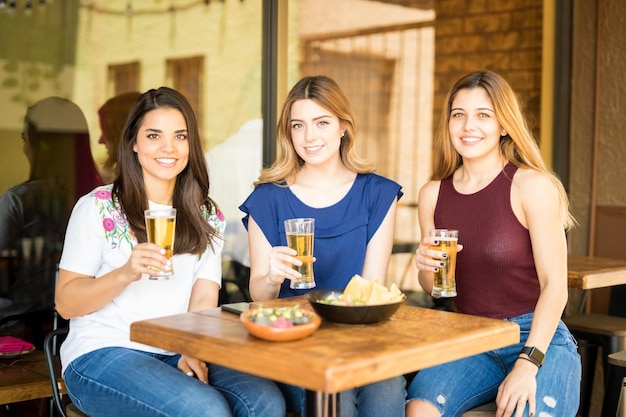 Retrato de um grupo de três amigas bonitas bebendo cervejas em um restaurante