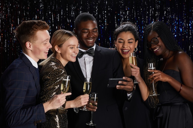 Retrato de um grupo multiétnico de amigos segurando taças de champanhe e transmitindo ao vivo online enquanto desfruta de uma festa elegante.