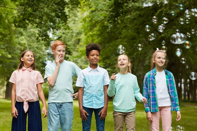Retrato de um grupo multiétnico de crianças em pé na fila ao ar livre enquanto brinca com bolhas no parque