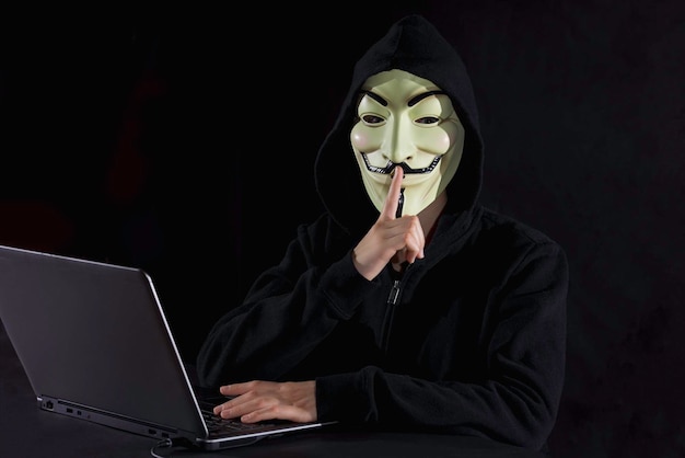 Retrato de um hacker de computador usando máscara com o dedo nos lábios contra um fundo preto
