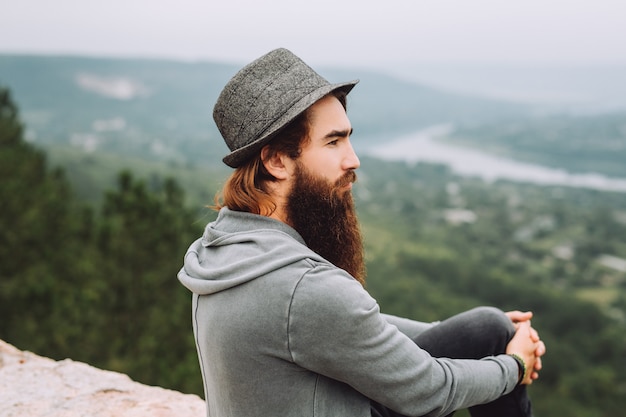 Retrato de um hippie barbudo elegante sério sentado em uma rocha em uma montanha no fundo de uma paisagem épica