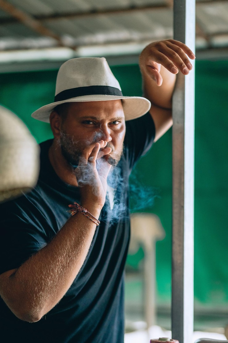 Foto retrato de um homem adulto brutal com um chapéu-panamá preto e branco com uma barba com um charuto na boca, fumando na rua. fundo verde.