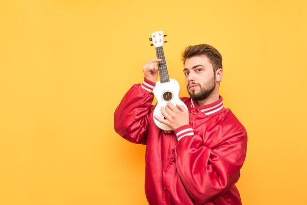 Foto retrato de um homem adulto com um ukulele nas mãos em amarelo e posando com uma jaqueta vermelha.