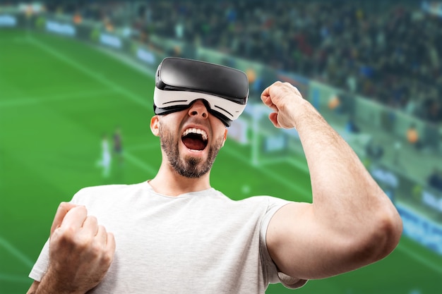 Retrato de um homem adulto feliz em óculos de realidade virtual, com a boca ligeiramente aberta, levantando a mão. O campo de futebol ao fundo está desfocado. O conceito de realidade virtual.