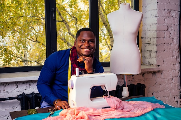 Retrato de um homem africano bonito sorridente costureira com máquina de costuraAfrio homem americano elegante designer trabalhando na oficina de alfaiate manequim fita métrica no quarto contra a janela de outono