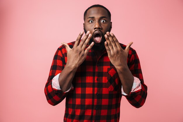 Retrato de um homem africano chocado, vestindo uma camisa xadrez, isolado na parede rosa, gesticulando