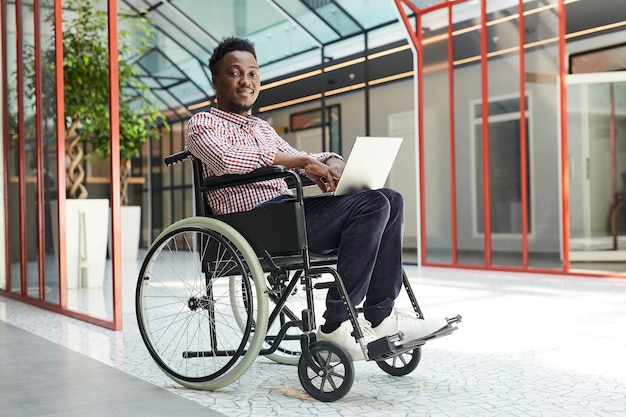 Retrato de um homem africano com deficiência sentado em uma cadeira de rodas e olhando enquanto trabalha no laptop