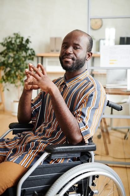 Retrato de um homem africano com deficiência, sentado em uma cadeira de rodas e olhando para a câmera, trabalhando no escritório