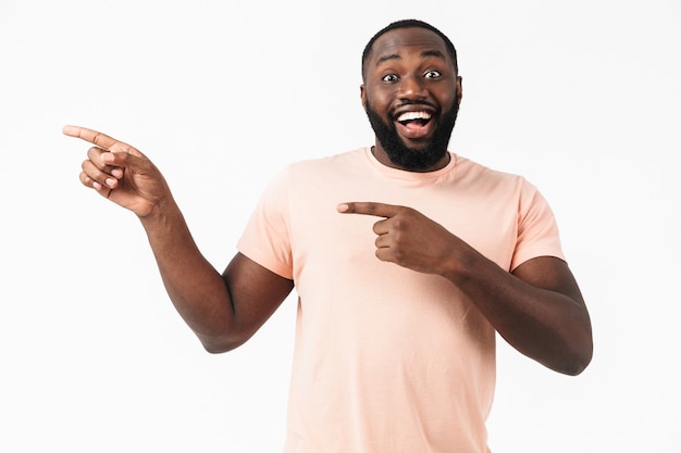 Retrato de um homem africano feliz e animado, vestindo uma camiseta em pé, isolado na parede branca, apontando para o espaço da cópia