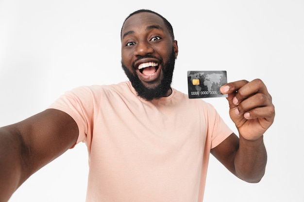 Retrato de um homem africano feliz vestindo camiseta em pé, isolado na parede branca, tomando selfie, mostrando o cartão de crédito