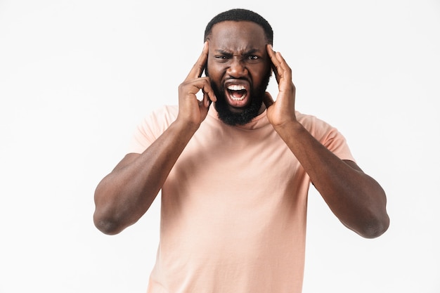 Retrato de um homem africano irritado, vestindo uma camiseta em pé, isolado na parede branca, com dor de cabeça