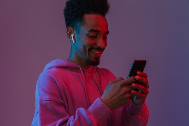 Retrato de um homem afro-americano barbudo com um capuz colorido falando ao celular com um fone de ouvido isolado sobre a parede violeta