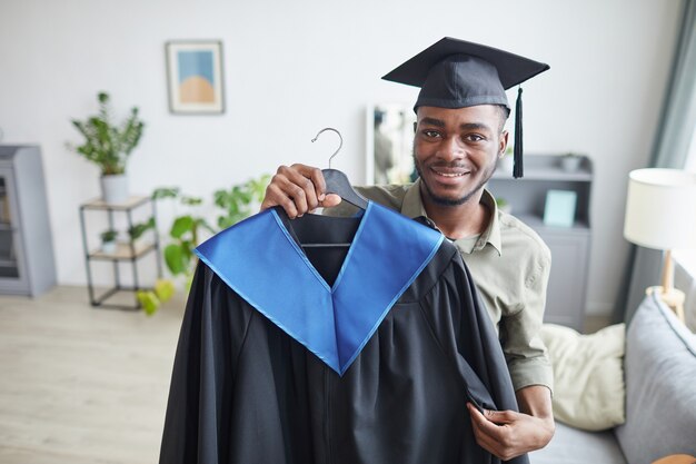 Retrato de um homem afro-americano feliz segurando um vestido de formatura e sorrindo para a câmera enquanto se prepara para a cerimônia em casa.