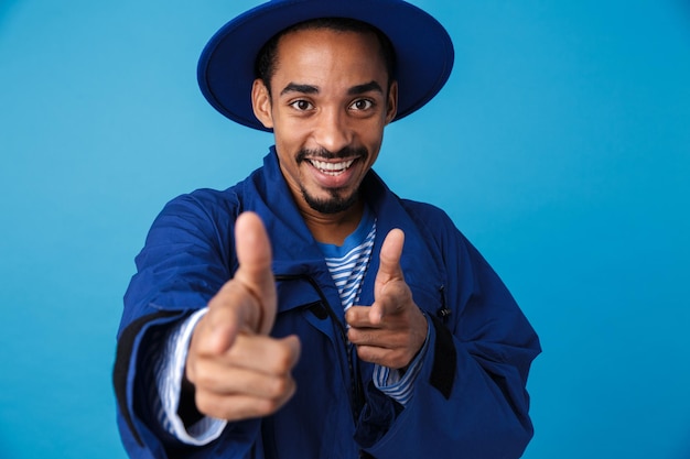 Retrato de um homem afro-americano feliz usando chapéu, sorrindo e apontando os dedos isolados no azul