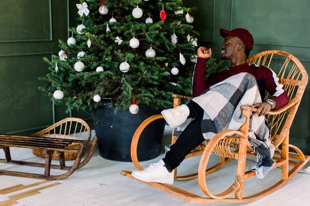 Retrato de um homem afro-americano hipster vestido com uma camisola de lã vermelha e boné vermelho, sentado em uma cadeira confortável perto da árvore de Natal